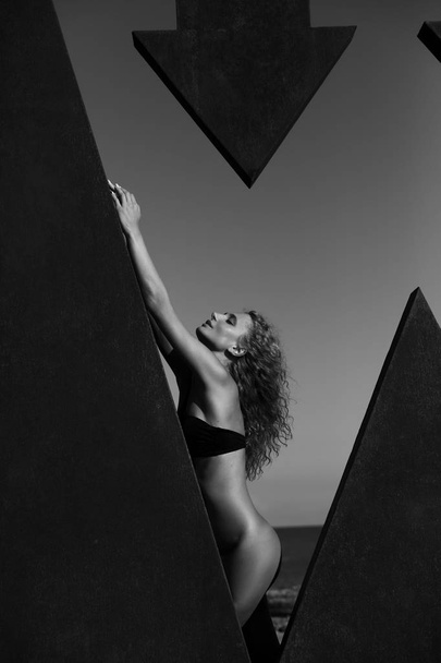 Sexy female model with curly hair weared in black bikini standing near sculpture at the sea. Black and white - Foto, afbeelding