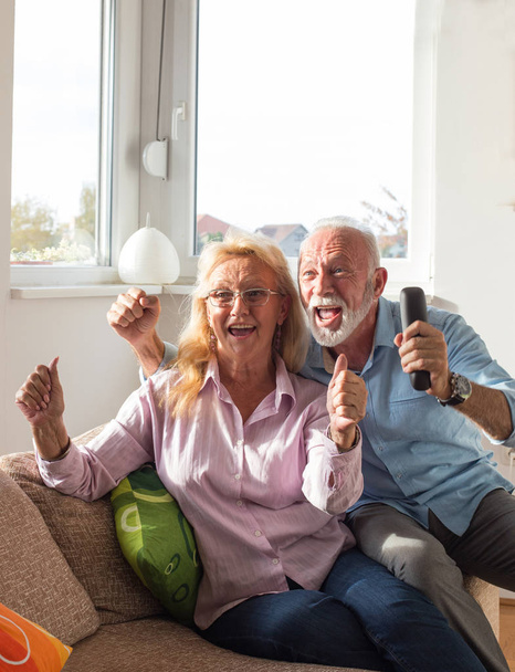 Aufgeregtes älteres Ehepaar schaut fern und jubelt auf Sofa im Wohnzimmer - Foto, Bild