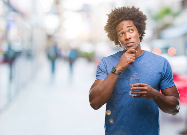 Afro american man drinking glass of water over isolated background serious face thinking about question, very confused idea - Zdjęcie, obraz