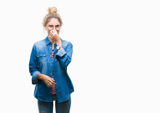 Young beautiful blonde woman wearing glasses over isolated background smelling something stinky and disgusting, intolerable smell, holding breath with fingers on nose. Bad smells concept. - Фото, изображение