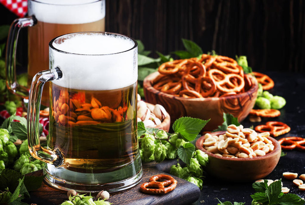 Light german beer poured into big glass, fresh green hops and bowls with salty snacks and nuts, autumn beer festival concept, dark background, selective focus - Photo, Image