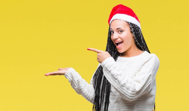 Плетені молодих афро-американських дівчина волосся капелюсі Різдво над ізольованих фон здивували і посміхається на камеру під час представлення рукою і вказуючи пальцем. - Фото, зображення