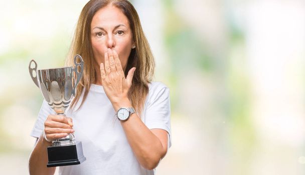Middelbare leeftijd hispanic winnaar vrouw vieren award trofee houden over geïsoleerde achtergrond dekking mond met de hand geschokt met schaamte voor fout, uitdrukking van angst, bang in stilte, geheime concept - Foto, afbeelding