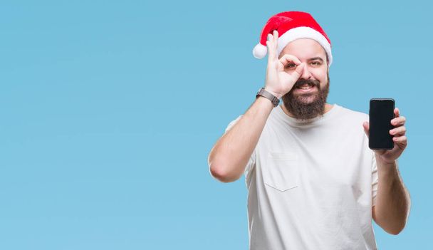 Young caucasian man wearing christmas hat showing smartphone screen over isolated background with happy face smiling doing ok sign with hand on eye looking through fingers - Valokuva, kuva