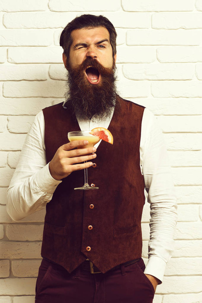 handsome bearded man with long beard and mustache has stylish hair on funny face holding glass of alcoholic beverage in vintage suede leather waistcoat on white brick wall studio background - Foto, afbeelding