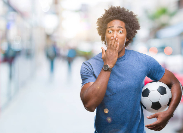 Αφρο αμερικανικό άνθρωπος που κρατά την μπάλα ποδοσφαίρου πάνω από το στόμα κάλυμμα απομονωθεί φόντο με το χέρι σοκαρισμένος με ντροπή για λάθος, έκφραση του φόβου, φοβάται στη σιωπή, μυστική έννοια - Φωτογραφία, εικόνα
