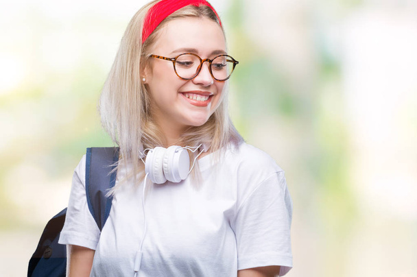 Jonge blonde student vrouw dragen van bril en rugzak over geïsoleerd achtergrond op zoek weg aan kant met glimlach op het gezicht, natuurlijke wijze van uitdrukking. Lachen vertrouwen. - Foto, afbeelding