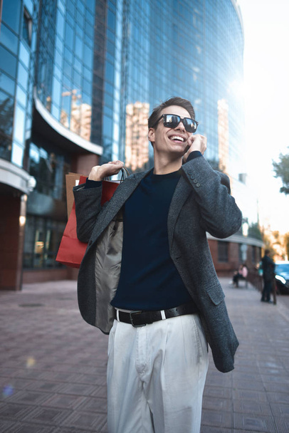 Positieve cool man met zonnebril en papieren zakken wandelen in de buurt van showcase met reflectie. - Foto, afbeelding