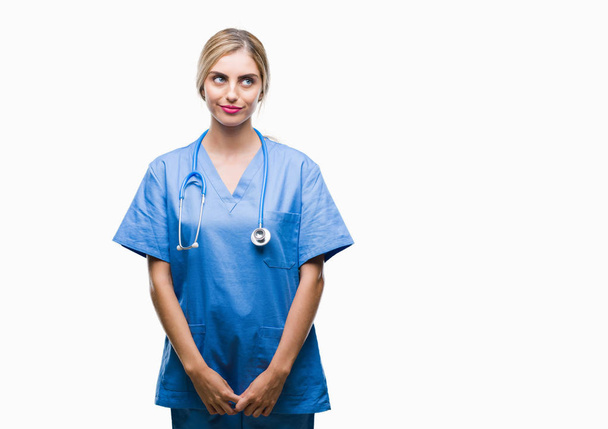 Молодая красивая блондинка врач хирург медсестра женщина на изолированном фоне улыбаясь, глядя в сторону и думая
. - Фото, изображение
