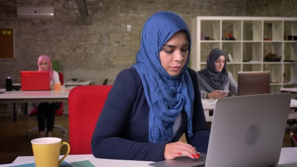 Yawning arabian woman in dark blue hijab is working on her laptop while other office females ias sitting behind, indoor illustration - Felvétel, videó