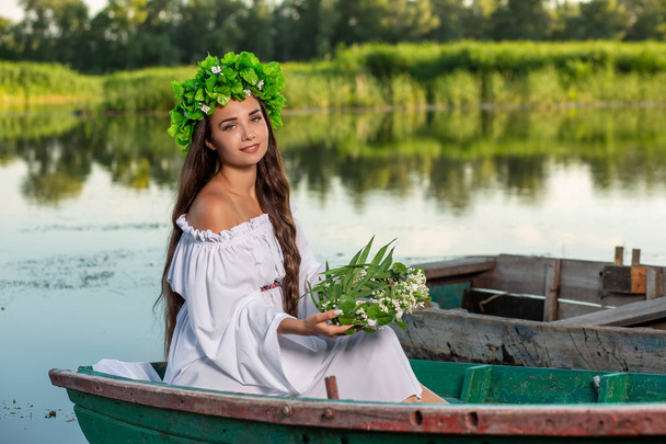De nimf met lang donker haar in een witte vintage jurk zitten in een boot in het midden van de rivier. - Foto, afbeelding