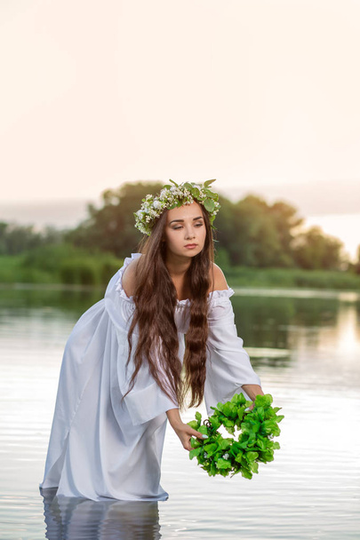 Beautiful woman on the river bank. Rustic woman with a wreath in a transparent dress - Photo, Image