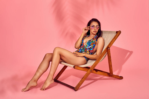 Zomer levensstijl mode portret van jonge prachtige vrouw op een lounge stoel, roze achtergrond, studio opname. Dragen van een stijlvolle zonnebril, bikini. - Foto, afbeelding
