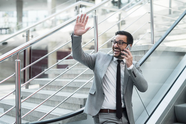 Empresario hablando en el teléfono inteligente y saludando al colega mientras sube las escaleras móviles
. - Foto, imagen