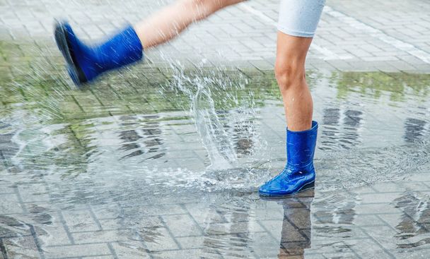 meisje in blauw rubber laarzen in een plas springen na een regen buiten op zomerdag. Close-up van de benen - Foto, afbeelding