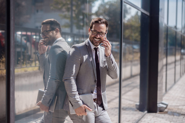 Портрет улыбающегося бизнесмена с помощью смартфона, опирающегося на окно. В другой руке планшет
. - Фото, изображение