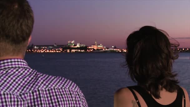 Man and woman standing on pier. Scene. Close-up from back of man and woman standing on pier. Evening view from pier to shining city on other side with sunset colored sky - Footage, Video