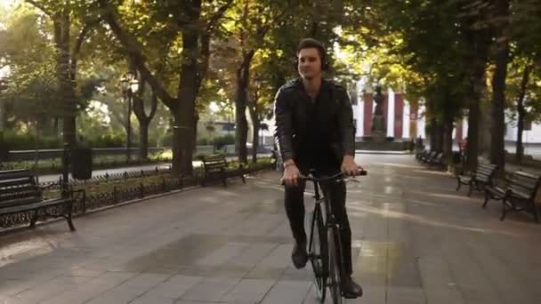 Young man with headphones riding bicycle in the city park on the trekking black bike. Riding and dancing while listening to the favourite music. Trees on the background. Front view - Materiał filmowy, wideo