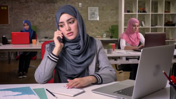 Confident unusual muslim girl in hijab has serious talk over phone while sitting, modern vibes, brick office, arabian women on background - Felvétel, videó