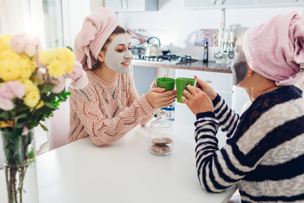Mutter und ihre erwachsene Tochter trinken Tee mit aufgesetzten Gesichtsmasken. Frauen chillen und reden in der Küche - Foto, Bild