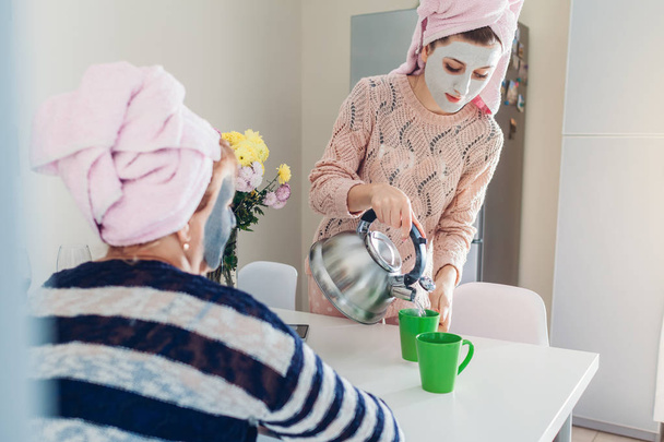 Young woman with her senior mother preparing tea with facial masks applied. Women chilling and talking on kitchen - Photo, Image
