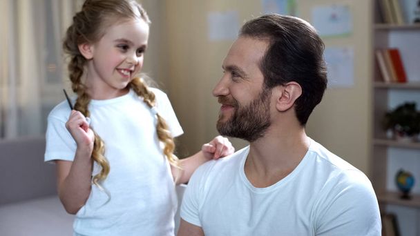 Little girl combing father hair and kissing cheek, playing games, family love - Фото, зображення