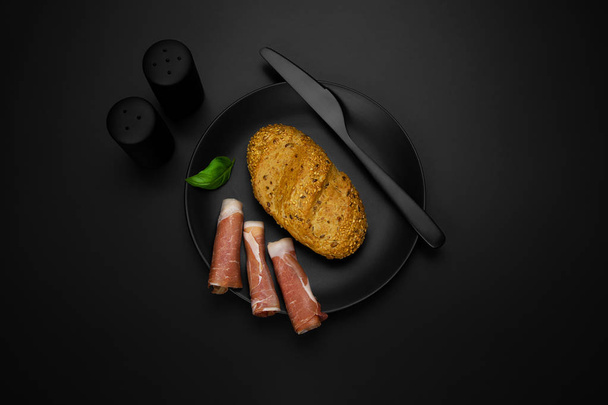 Prosciutto ham with roll and basil on a black plate, - Photo, Image