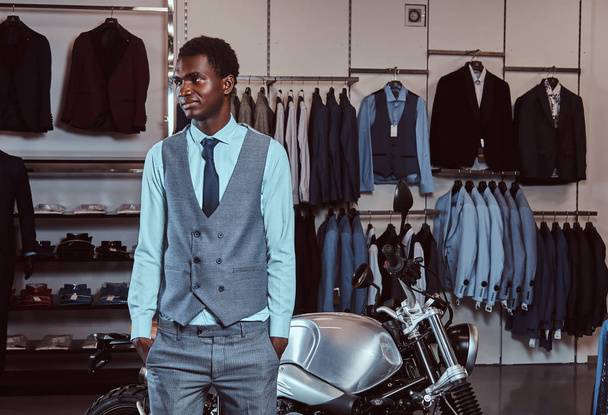 Elegantemente vestito afroamericano giovane in posa vicino a moto sportive retrò presso il negozio di abbigliamento uomo
. - Foto, immagini