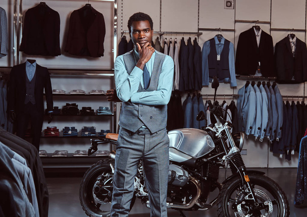 Elegantemente vestido joven afroamericano posando cerca de moto deportiva retro en la tienda de ropa para hombres
. - Foto, Imagen