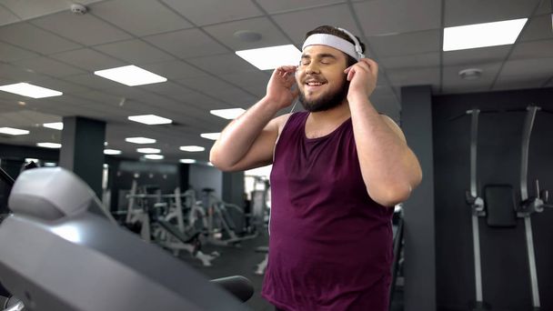 Fat man putting on headset and listening to music during jogging on treadmill - Zdjęcie, obraz