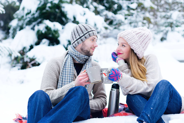 para zakochanych w ciepłe ubrania siedzieć w śniegu i przytrzymaj kubki z kubkiem gorącego napoju i spojrzeć na siebie - Zdjęcie, obraz
