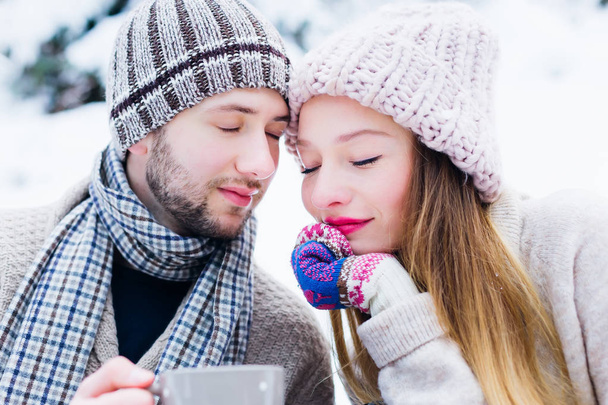 the couple in love leaned against each other and closed their eyes. they hold the cups in their hands and sit on the snow in warm clothes - Photo, Image
