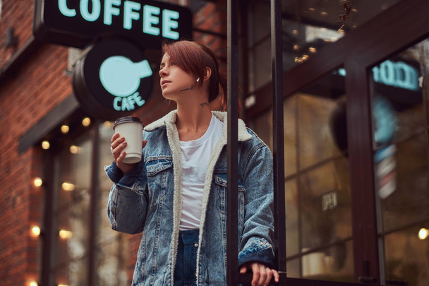 A beautiful girl wearing a denim coat holding cup with takeaway coffee outside near the cafe. - Photo, Image