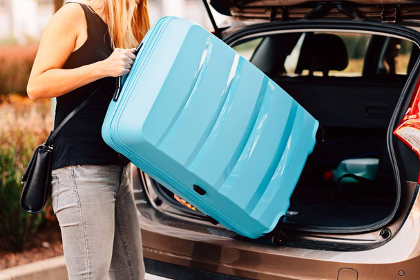 Young woman loading two blue plastic suitcases to car trunk. Preparation for trip - Zdjęcie, obraz