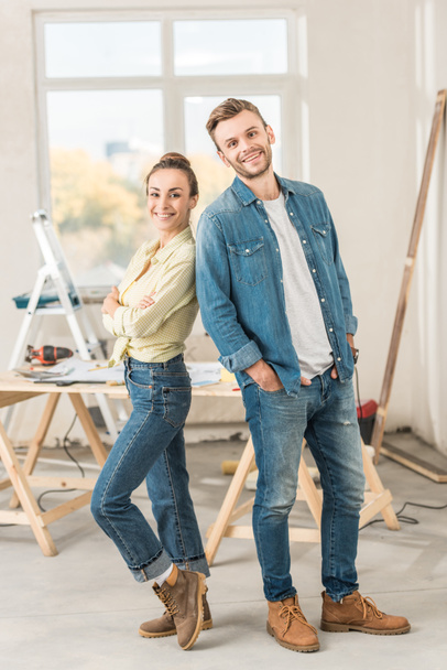 onnellinen nuori pari seisoo selkä selkää vasten ja hymyilee kameralle talon korjauksen aikana
 - Valokuva, kuva