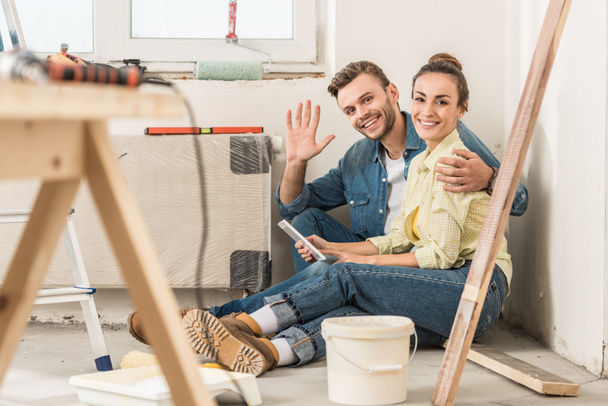 happy young couple using smartphone and smiling at camera while sitting on floor in new house - Photo, Image