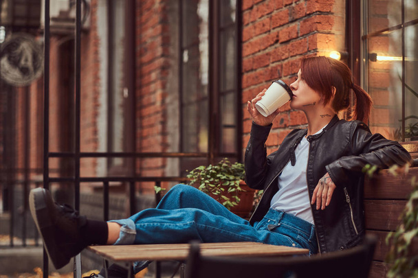Redhead girl wearing trendy clothes sitting outside the cafe and drinks takeaway coffee. - 写真・画像