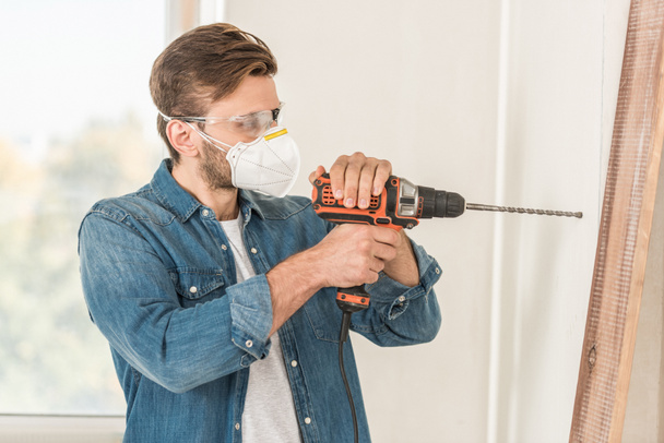 young man in protective mask and goggles using electric drill during house repair - Photo, Image