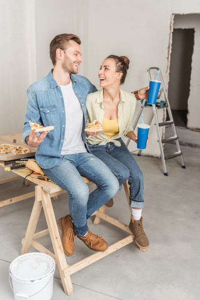 glückliches junges Paar mit Pizza und Pappbechern, die zusammen sitzen und einander während der Reparatur lächeln - Foto, Bild