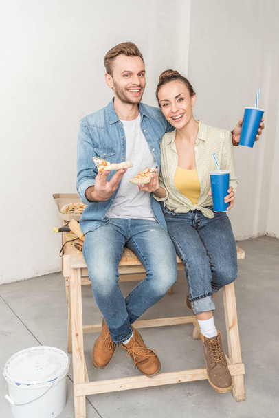 feliz jovem casal com copos de papel e fatias de pizza sentados juntos e sorrindo para a câmera no novo apartamento
 - Foto, Imagem