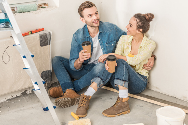 vysoký úhel pohledu usměvavý mladý pár drží papírových kelímků a sedí na podlaze v novém bytě - Fotografie, Obrázek