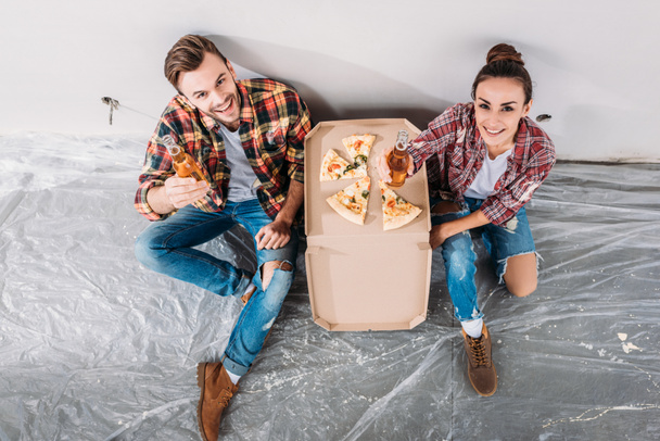 vista aérea de feliz pareja joven con cerveza y pizza sentado en el suelo y sonriendo a la cámara en el nuevo apartamento
 - Foto, imagen