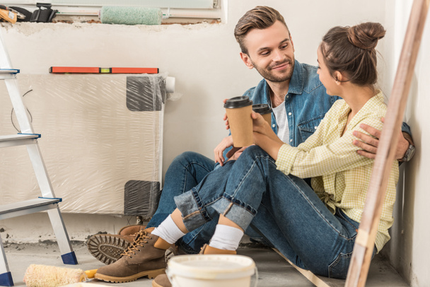 young couple holding paper cups and smiling each other while sitting on floor during house repair - Photo, Image