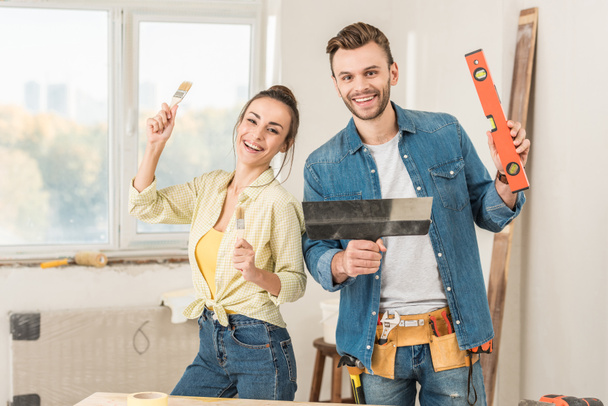 happy young couple holding tools and smiling at camera during house repair - Photo, Image