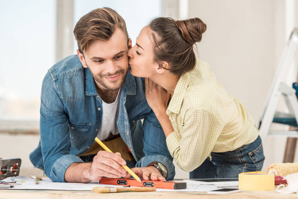 young woman kissing smiling man marking blueprint with level tool  - Foto, imagen