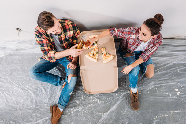 overhead view of young couple clinking beer bottles and eating pizza while sitting on floor in new apartment - Photo, Image