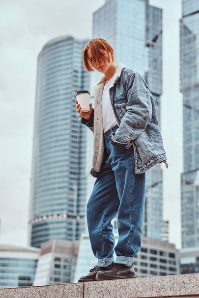 Stylish redhead hipster girl with tattoo on her face wearing denim jacket holding takeaway coffee in front of skyscrapers in Moskow city. - Photo, Image