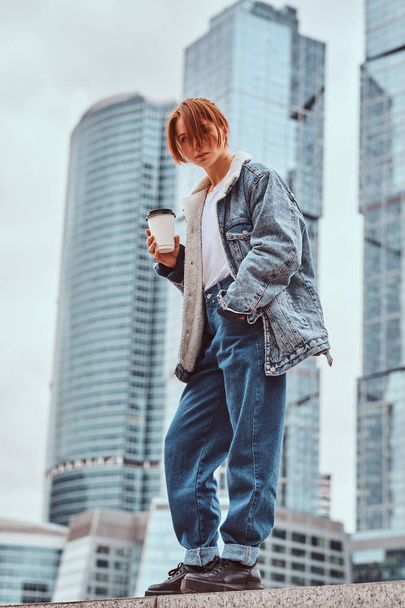 Стильная рыжая хипстерша с татуировкой на лице в джинсовой куртке с кофе на вынос перед небоскребами в Москве
. - Фото, изображение