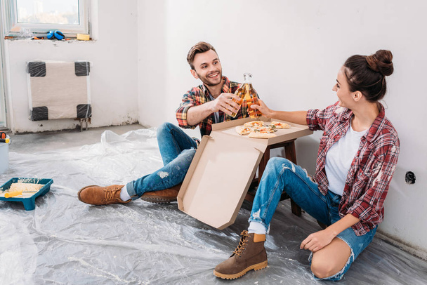 onnellinen nuori pari clinking olutpulloja ja syö pizzaa istuessaan lattialla uudessa asunnossa
 - Valokuva, kuva