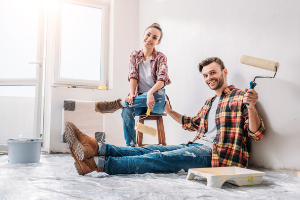 happy young couple holding paint rollers and smiling at camera during renovation - Fotoğraf, Görsel